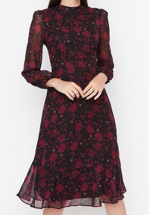 Kobieta ubrana w czarną sukienkę z długim rękawem z czerwonym kwiatowym nadrukiem, z wysokim dekoltem i falbaną na dole, z rękami luźno złożonymi na wysokości talii.