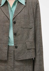 Bruine geruite cropped jas met een enkele borstzak, drie knopen en een contrasterend lichtblauw overhemd eronder.