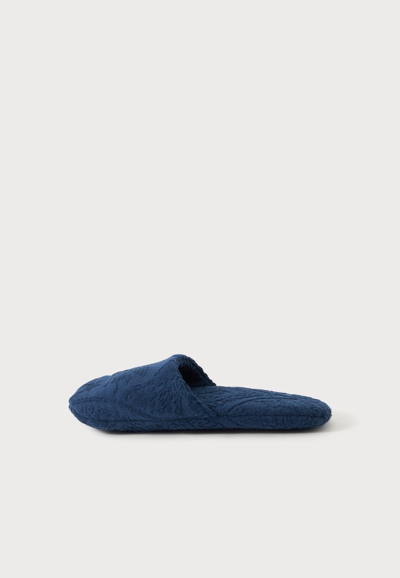 Une pantoufle à enfiler bleu marine unique avec un tissu texturé et un dos ouvert, vue de côté sur un fond clair uni.