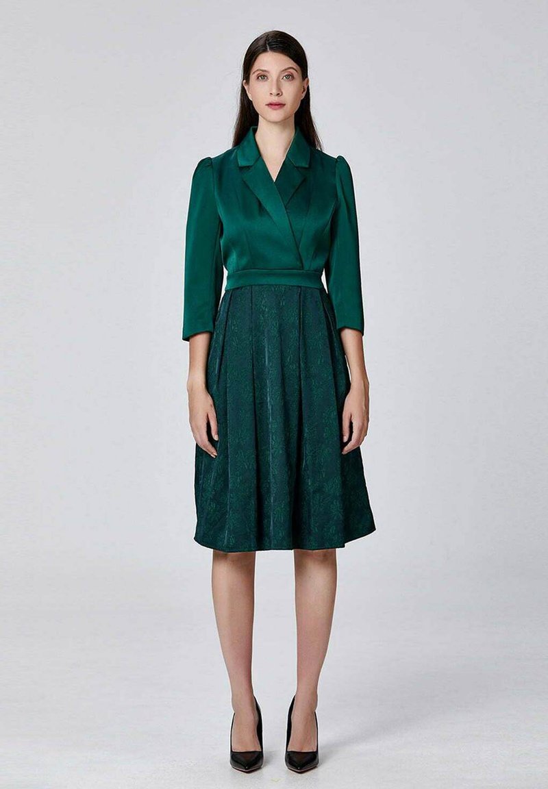 Femme portant une robe verte foncée, longueur genou, avec une jupe texturée et des manches trois-quarts, se tenant devant un fond clair.
