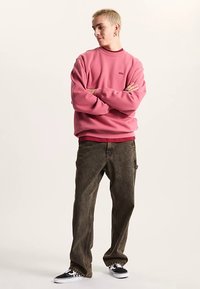 Różowa bluza z prążkowanymi mankietami i małym logo, zestawiona z luźnymi, ciemnobrązowymi spodniami typu cargo oraz czarnymi trampkami z białymi podeszwami.