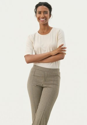 Donna sorridente con capelli scuri raccolti, indossa una maglietta a maniche corte color crema e pantaloni beige a quadri, in piedi con le braccia incrociate davanti a uno sfondo semplice.
