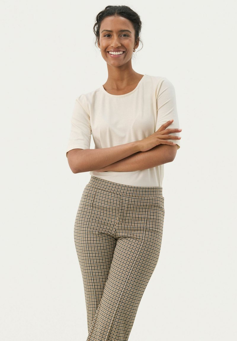 Femme souriante aux cheveux foncés attachés, vêtue d'un haut crème à manches courtes et d'un pantalon beige à carreaux, debout les bras croisés devant un fond uni.