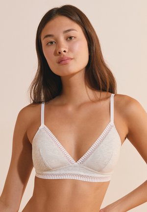 Joven mujer con cabello castaño largo que lleva un bralette gris claro con tirantes blancos finos sobre un fondo beige.