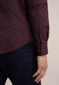 Burgundy skjorta med en slät textur, med knäppningsmanschetter och rundad nederkant, tillsammans med mörkblå jeans med en bakficka.