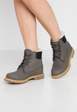 Nogi w szarych zimowych butach na sznurowadła z beżowymi podeszwami i białym futrzanym wykończeniem, stojące na jasnoszarej podłodze na jednolitym tle.