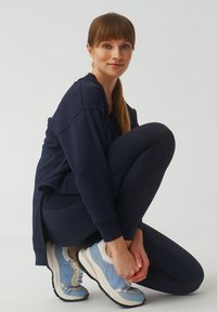 Marinesweater met geribbelde textuur, gecombineerd met donkere legging. Blauwe sportschoenen hebben mesh panelen en een forse zool.