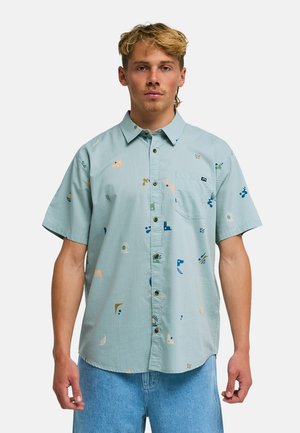 Młody mężczyzna z krótkimi blond włosami, ubrany w błękitną koszulę z krótkim rękawem, zapinaną na guziki, zdobioną kolorowymi abstrakcyjnymi kształtami oraz jasnoniebieskie dżinsy.