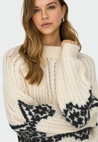 Crèmewitte gebreide trui met een losse pasvorm, brede manchetten en een contrasterend zwart geometrisch patroon op de mouwen. Details van gestructureerd garen zijn zichtbaar.