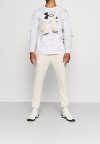 Langärmliges graues Camouflage-Shirt mit schwarzem Logo, beige Hose und weißen Sportschuhen. Pullover über der Schulter getragen.