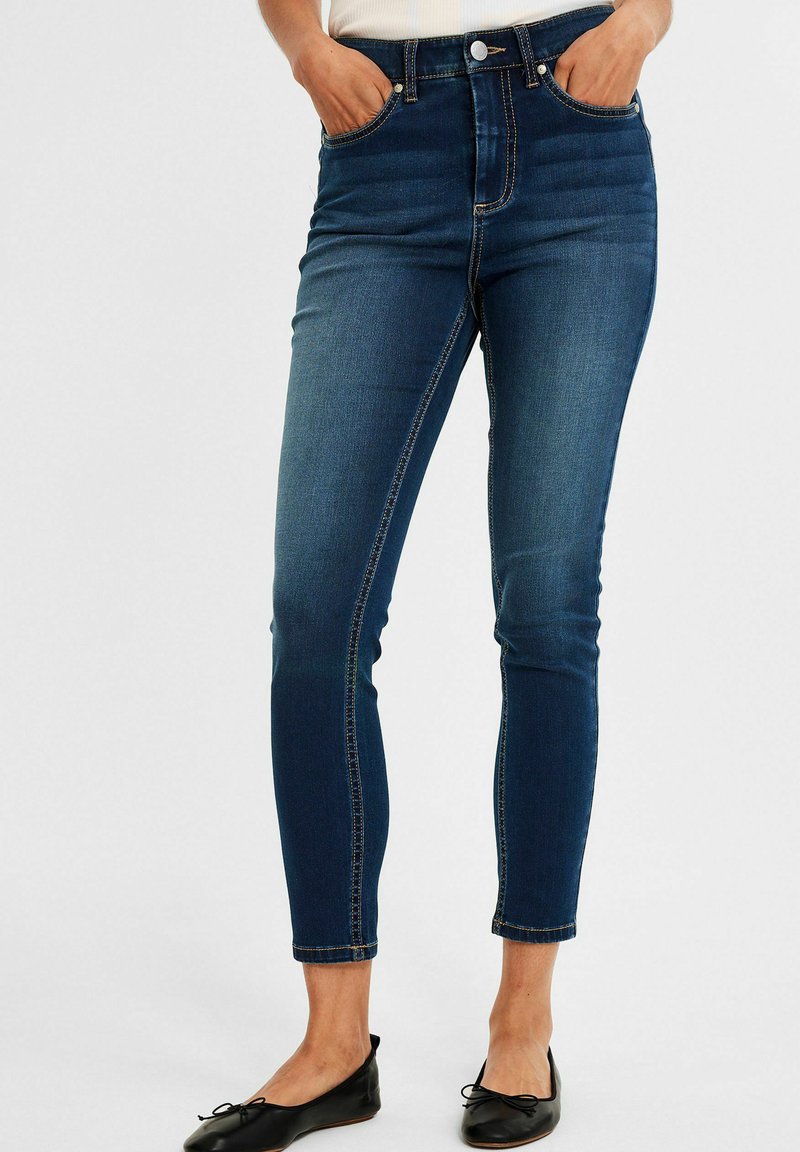 Jeansy z wysokim stanem w ciemnoniebieskim kolorze, o dopasowanym kroju, z przednimi kieszeniami, kontrastowym przeszyciem i podwiniętym dołem.