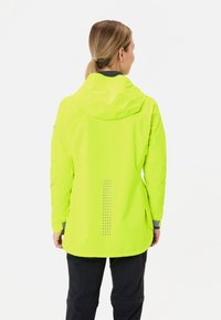Vrouw met blond haar in een paardenstaart, draagt een heldergele regenjas en zwarte broek, gezien vanaf de achterkant op een witte achtergrond.