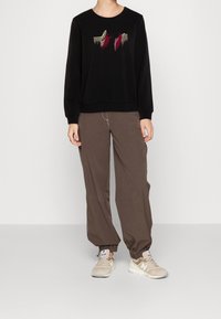 Sudadera negra con diseño bordado en rojo y dorado, combinada con pantalones marrones holgados y zapatillas color beige con suelas blancas.