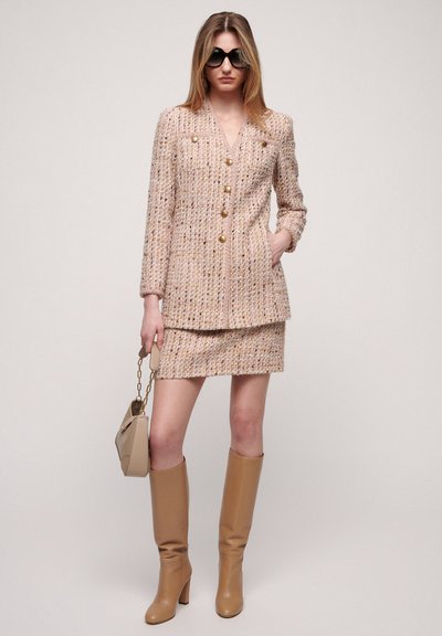 Tweed blazer and mini skirt set in pastel pink, with gold button accents. Worn with tall beige boots and a small beige handbag.