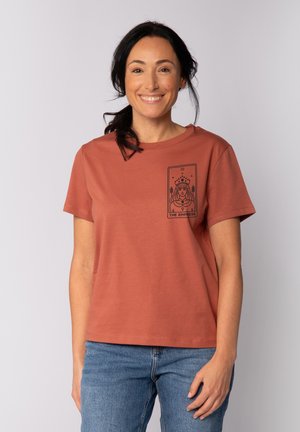 T-shirt en coton couleur rouille à manches courtes et coupe décontractée, avec un imprimé graphique noir d'une femme sur le côté gauche de la poitrine.