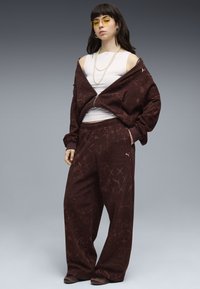 Femme portant un survêtement marron à motifs oversize, un haut sans manches blanc, des lunettes à verre jaune, un collier de perles et des chaussures marron, se tenant devant un fond gris.