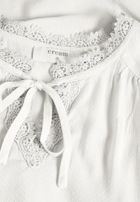Blusa color crema con un tessuto texturizzato, caratterizzata da un colletto rifinito in pizzo e un fiocco sul davanti. L'etichetta è visibile.