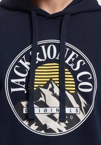 Dunkelblaues Hoodie mit einem runden Logo, das Berge, eine gelbe Sonne mit Streifen und den Text "JACK & JONES CO ORIGINALS" in Weiß zeigt.