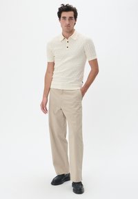 Kötött krém színű polo ing texturált mintával, h�árom gombos gallérral és rövid ujjakkal, bézs nadrággal és fekete cipővel párosítva.