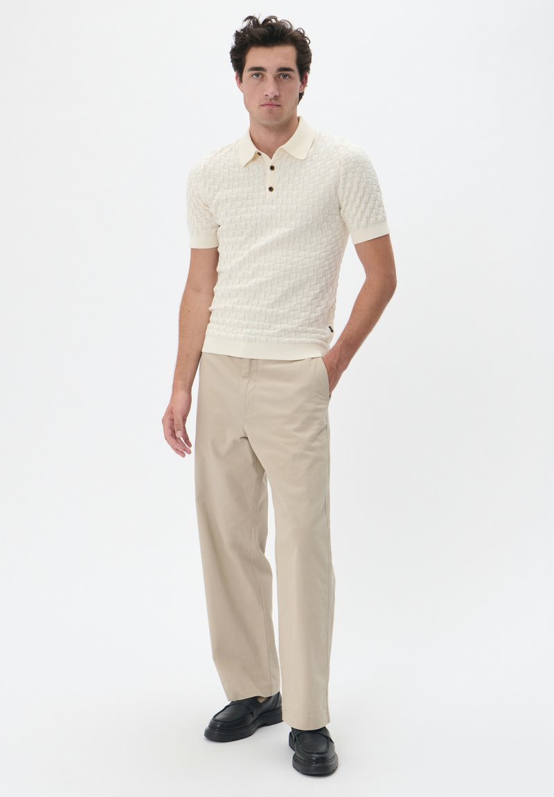 Kötött krém színű polo ing texturált mintával, három gombos gallérral és rövid ujjakkal, bézs nadrággal és fekete cipővel párosítva.