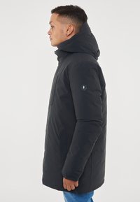 Giacca a vento nera con cappuccio. Presenta una zip frontale, tasche laterali e un patch con logo sul maniche sinistro. Tessuto liscio e isolante.