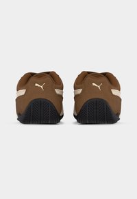 Ζευγάρι καφετιών και μπεζ αθλητικών παπουτσιών Puma, φαίνεται από πίσω, με έμφαση στις μαύρες λαστιχένιες σόλες και το λογότυπο Puma στη φτέρνα.