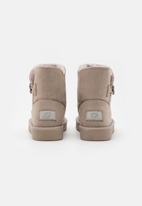 Bottines en daim tan avec doublure en peluche, bords arrondis et décorations en métal argenté. Présente un petit logo en forme de cœur sur un patch gris.