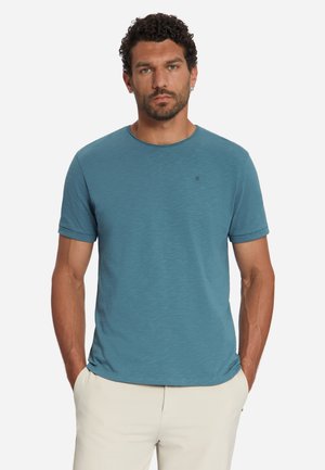 Mann mit kurzen lockigen Haaren und Bart, trägt ein türkisfarbenes T-Shirt mit kurzen Ärmeln und hellbeige Hose, steht mit Händen in den Taschen.