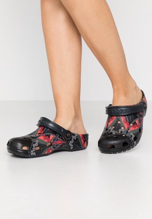 Beine mit schwarzen Crocs, die rote und graue abstrakte Muster aufweisen, stehend auf einem glatten weißen Boden vor einem einfarbigen hellen Hintergrund.