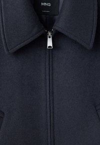 Veste zippée bleu marine foncé avec un col, comportant une fermeture éclair argentée visible et une étiquette de marque MNG à l’intérieur du col.