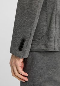 Grauer Blazer aus strukturiertem Stoff, mit einer einzelnen Reihe von vier schwarzen Knöpfen am Ärmel und einer Seitentasche.