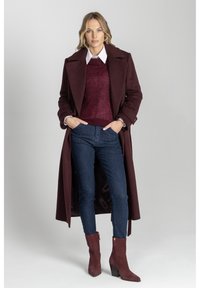 Kobieta w długim burgundowym płaszczu, dopasowanej bluzie i butach, białej koszuli z kołnierzykiem oraz ciemnoniebieskich, dopasowanych dżinsach, stojąca z rękami w kieszeniach.