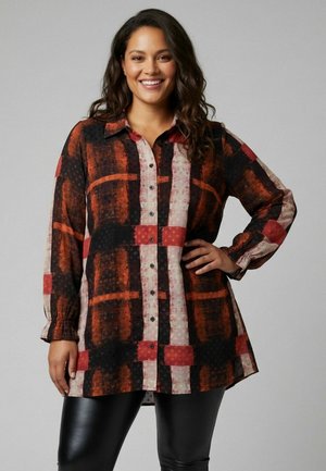 Femme souriante portant une chemise à manches longues à carreaux rouges, noirs et beiges, et un pantalon en cuir noir, posant avec la main sur la hanche.