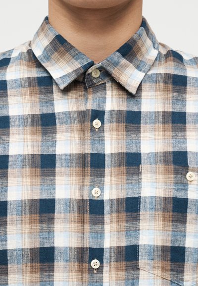 Κοντινό πλάνο ενός ατόμου που φοράει καρό πουκάμισο με κουμπιά, με μπλε, καφέ και λευκό μοτίβο σε τετράγωνα και μία τσέπη στο στήθος.