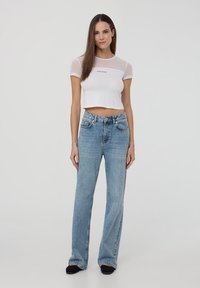 T-shirt bianco corto con maniche corte trasparenti, caratterizzato da un dettaglio testuale nero. Jeans azzurri a palazzo con finitura consumata e tasche.
