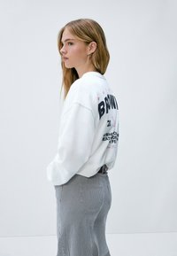 Sudadera blanca de corte oversize, con un gráfico en rosa y negro en la parte posterior, combinada con pantalones a rayas grises. Tejido suave, escote relajado.