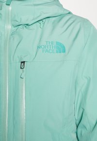 Helle Teal-Jacke mit Kapuze, ausgestattet mit einem Frontreißverschluss und einem markanten Logo auf der Brust. Hergestellt aus glattem, wetterfestem Material.