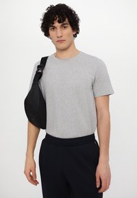 Čierna crossbody taška vyrobená z hladkého materiálu, s zakriveným tvarom a nastaviteľným popruhom. Nosená cez svetlosivý tričko s okrúhlym výstrihom.