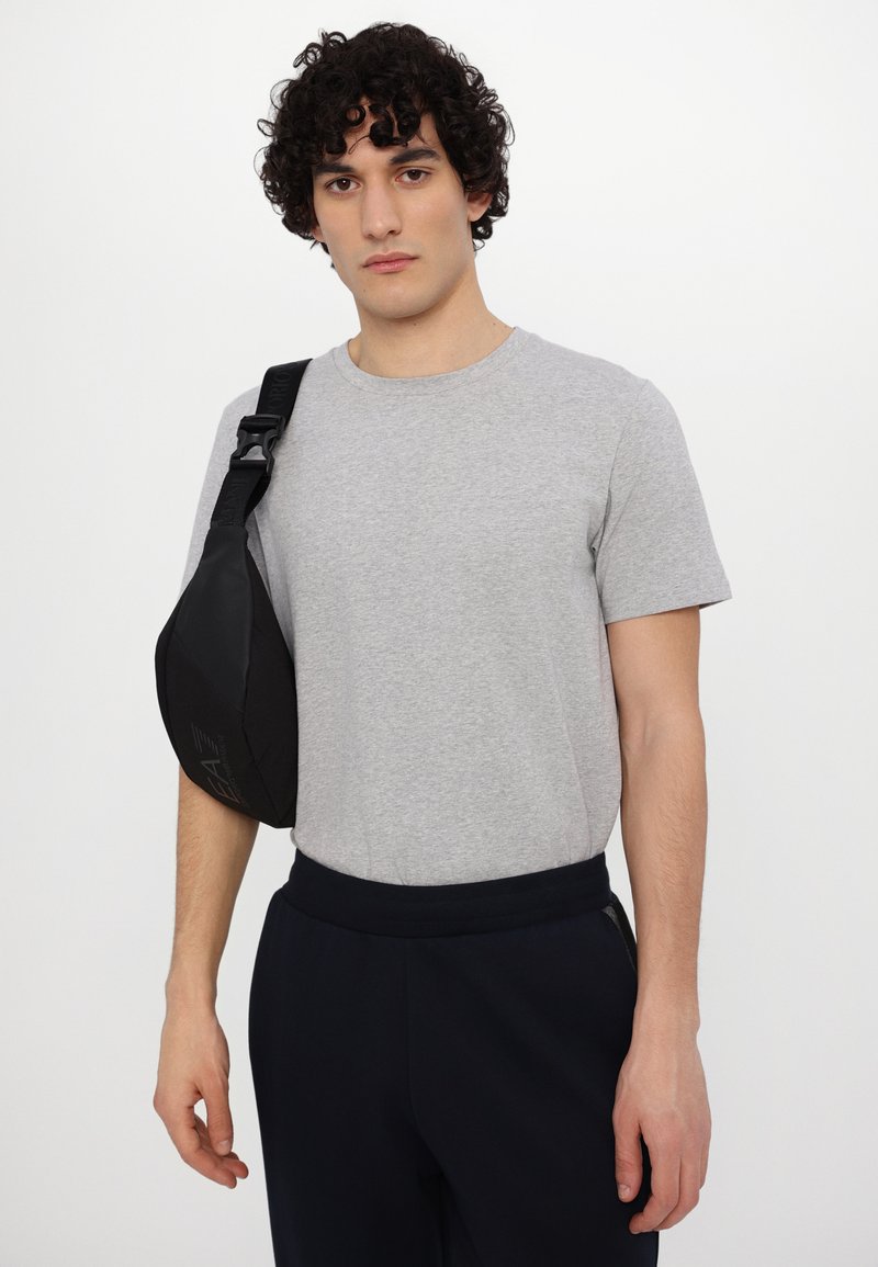 Borsa a tracolla nera realizzata in materiale liscio, con forma curva e tracolla regolabile. Indossata sopra una t-shirt a girocollo grigio chiaro.