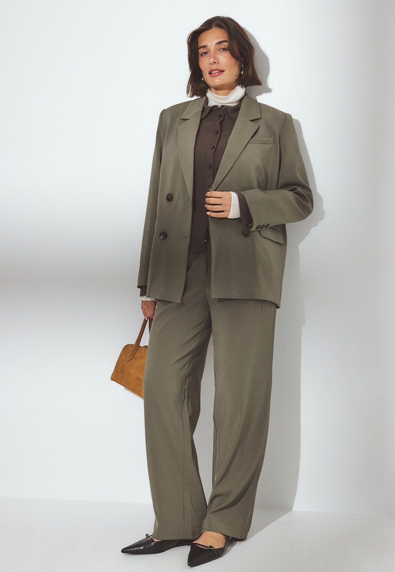 Ženska v olivno zeleni oversized obleki z rjavim puloverjem, belim rolko in črnimi špičastimi balerinkami, v roki drži rjav nahrbtnik.