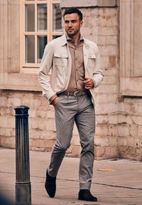 Lichtbeige jas met twee voorkantenzakken, een bruine shirt, grijs geruite broek en donkerbruine enkelboots. Achtergrond van een poreuze stenen muur.