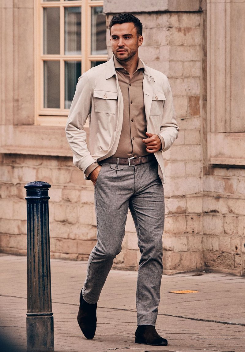 Lichtbeige jas met twee voorkantenzakken, een bruine shirt, grijs geruite broek en donkerbruine enkelboots. Achtergrond van een poreuze stenen muur.