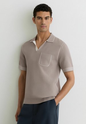 Młody mężczyzna ubrany w taupe krótki sweter polo z białym przeszyciem i granatowe spodnie, stojący na tle białej ściany.