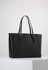 Černá taška přes rameno vyrobená z texturovaného syntetického materiálu, s dvěma dlouhými popruhy a minimalními akcenty ve zlatém tónu.