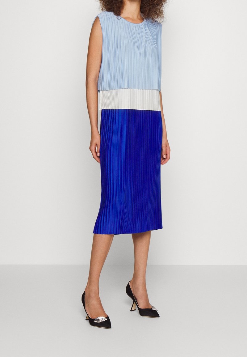 Femme portant une robe midi sans manches plissée à blocs de couleurs bleu clair, blanc et bleu roi avec des escarpins noirs pointus.
