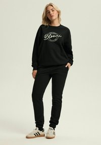 Czarna bluza z białym napisem, luźny krój, ściągacze na rękawach, zestawiona z pasującymi czarnymi dresami i białymi sneakersami z czarnymi paskami.