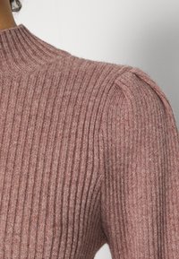Pull à maille côtelée de couleur mauve douce, présentant un col montant et un détail structuré aux épaules, mettant en valeur un tissu texturé.