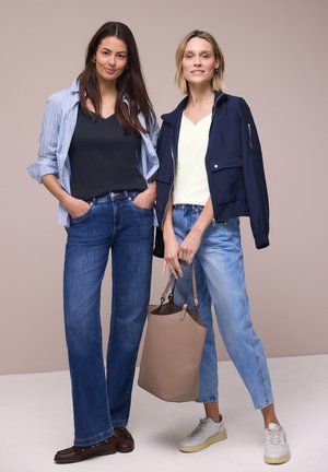 Due donne in piedi, indossano jeans e giacche casual; una tiene una borsa tote beige, entrambe sorridono davanti a uno sfondo beige uniforme.