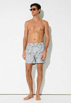 Mees, kes kannab tumedaid päikeseprille ja halli ujumispükst valgete lillemustritega, seisab paljajalu valge müüri vastu.