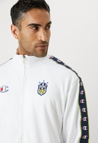 Witte zip-up jas met hoge kraag, voorzien van een blauw en geel gestreept zijpatroon, logo op de borst en een gladde textuur.